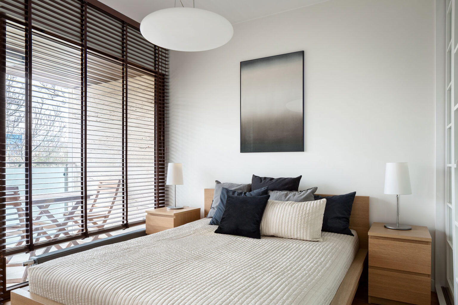 Big and comfortable bed in bright bedroom with big window with wooden blinds