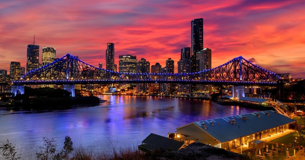 A vibrant sunset over a city skyline, featuring the Story Bridge illuminated in bright purple and blue lights.
