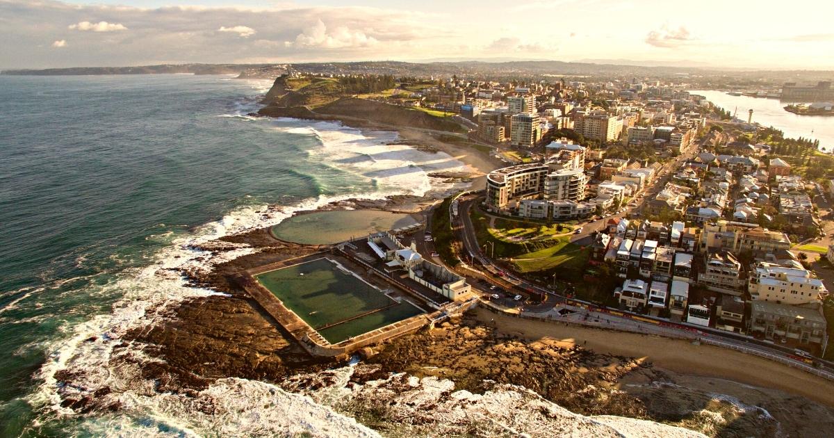 An aerial view of a coastal city at golden hour, featuring a large ocean bath and rock pool built directly into the rugged shoreline.