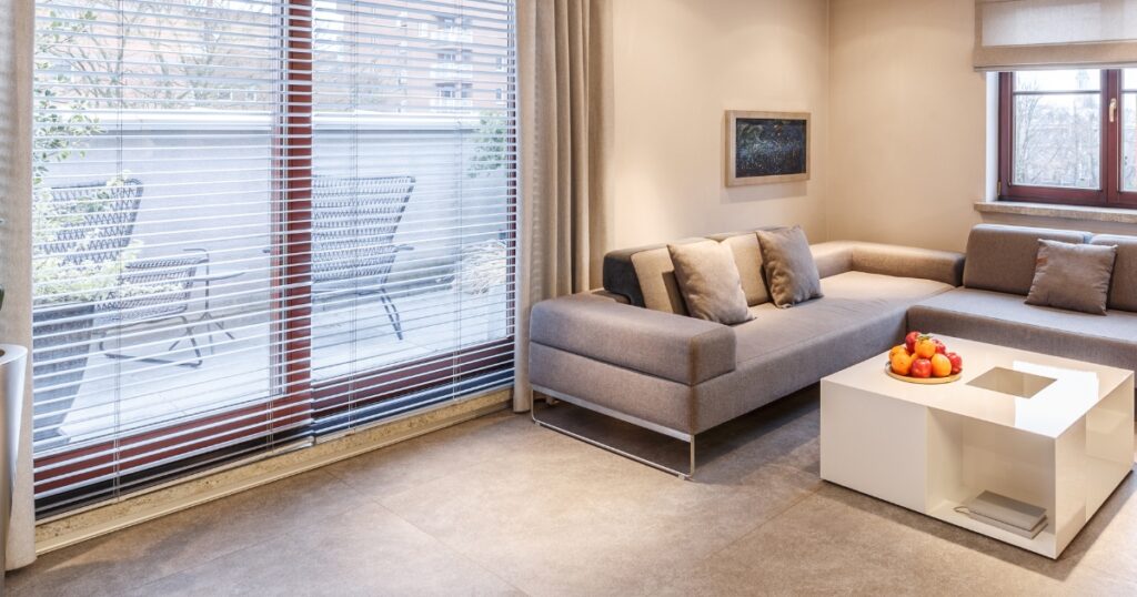 A modern, neutral-toned living room featuring a grey sectional sofa and a white geometric coffee table with a bowl of fruit.