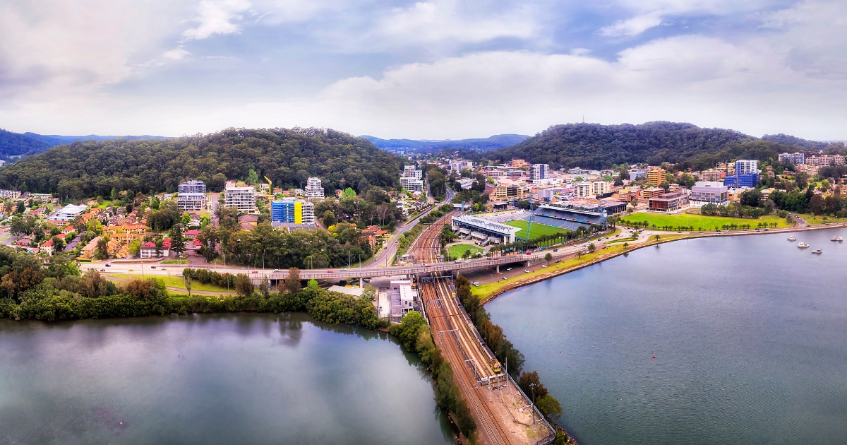 An aerial panoramic view of a coastal town featuring a railway line and a bridge crossing over a calm body of water.