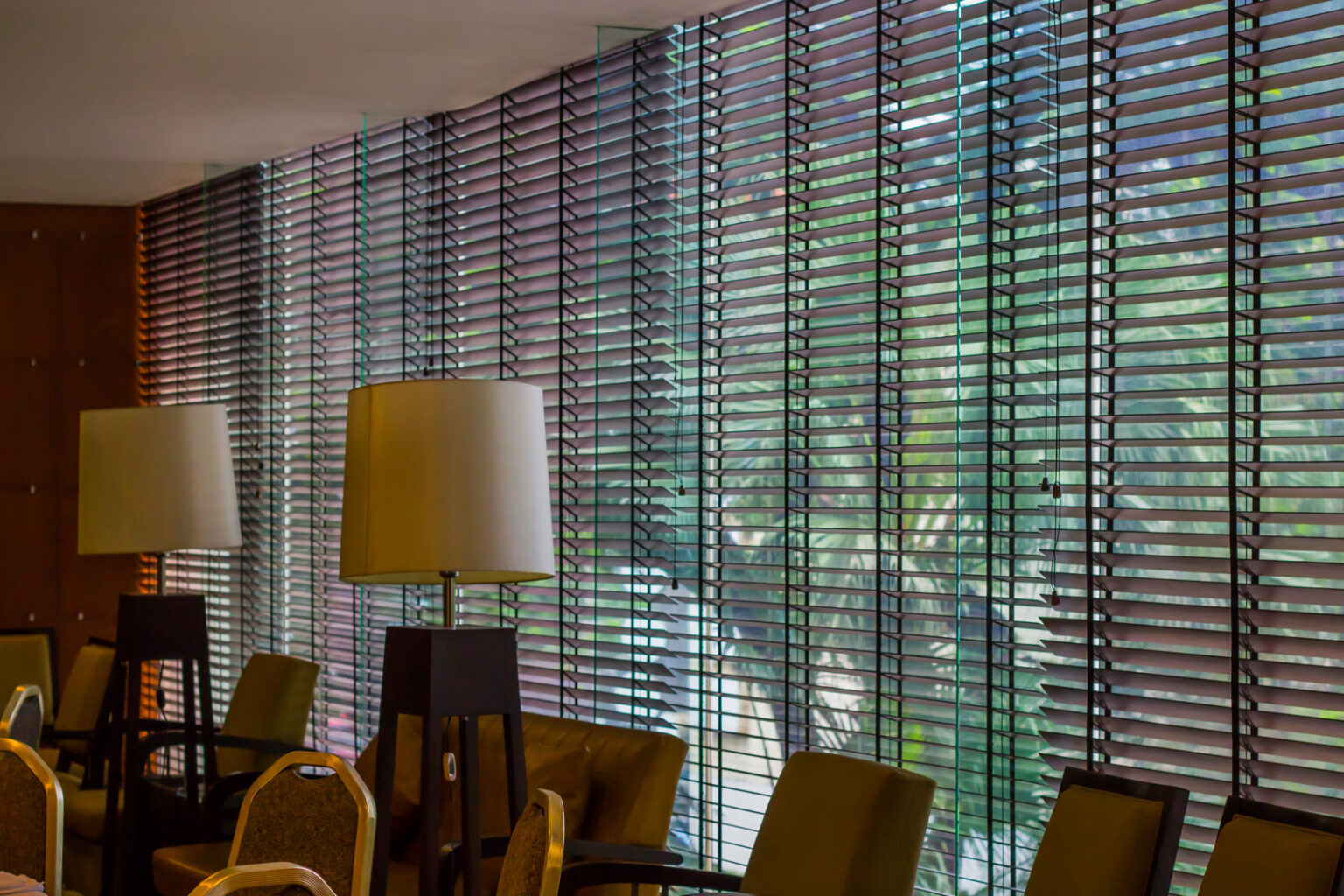 A dark horizontal venetian blinds covering a large window that overlooks tropical green foliage.