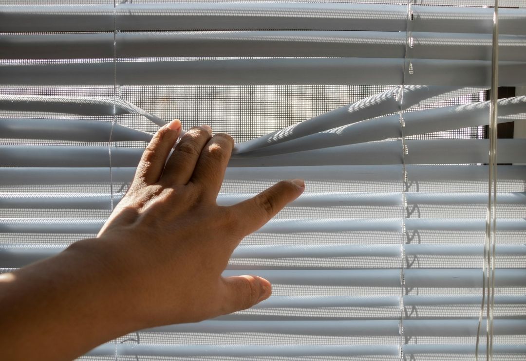 A close-up of a person's hand reaching in and pulling up a section of light grey or white horizontal blinds to peek out the window.