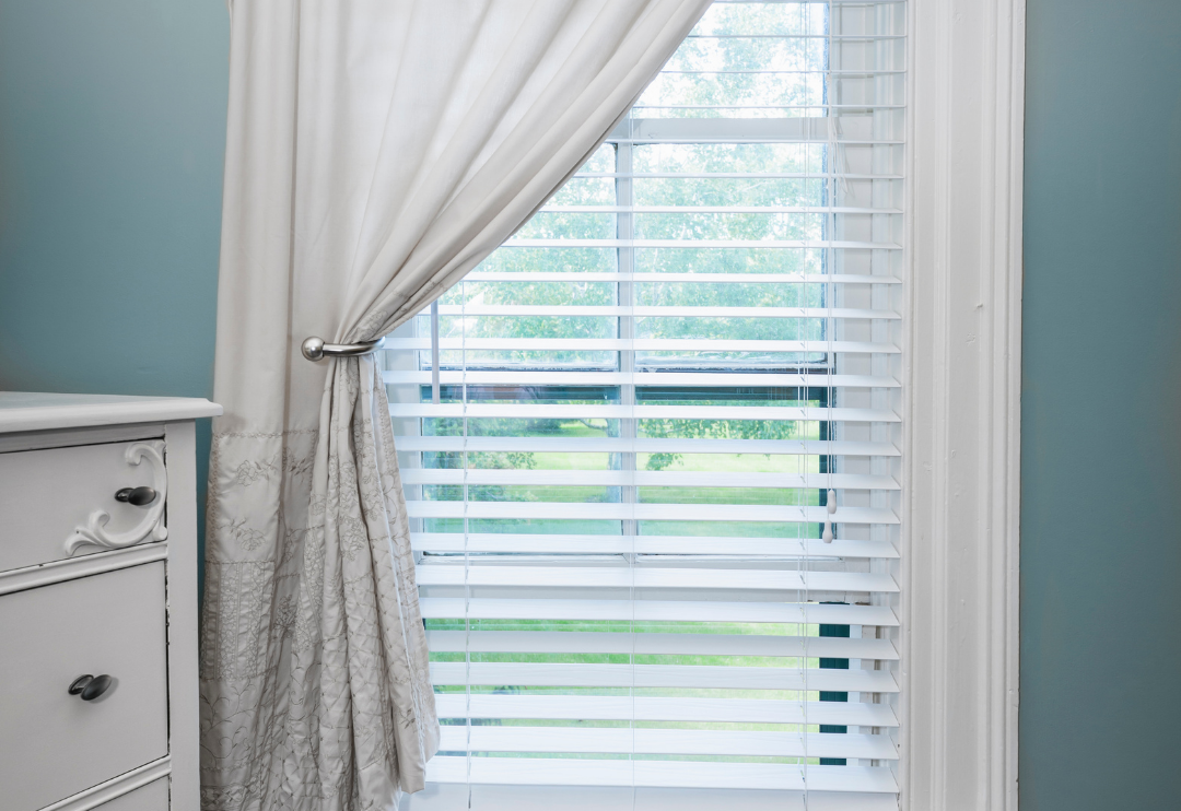 A set of white horizontal blinds covers a window, with the slats slightly open to reveal a green outdoor scene.