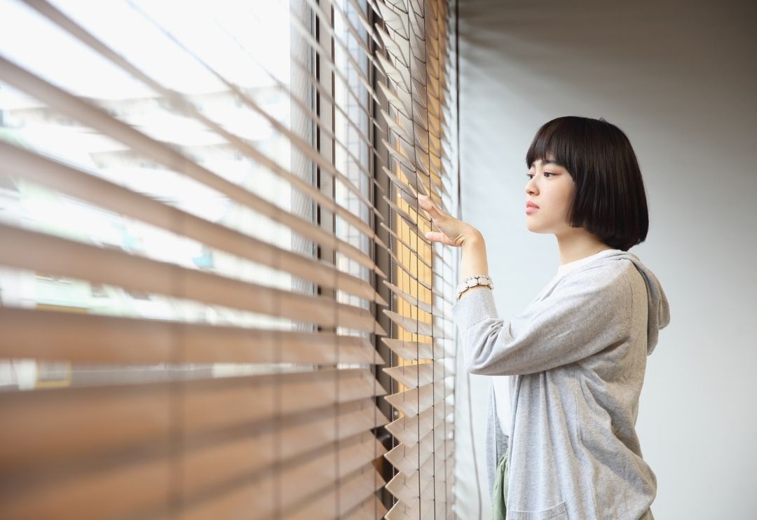 A woman with short dark hair, wearing a light grey hoodie, stands next to a large window with horizontal wooden blinds.