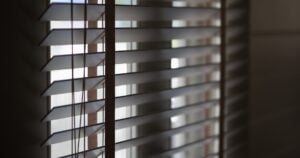A close-up, moody view of wooden Venetian blinds with dark decorative tapes, partially tilted to reveal bright light coming through the window.