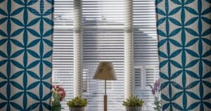 A window featuring white horizontal blinds framed by blue and white geometric patterned curtains