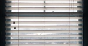 A close-up shot of white horizontal Venetian blinds partially closed over a window, with the pull strings hanging down on the left.