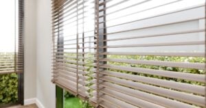 A close-up, angled view of light-coloured wooden Venetian blinds on a window, offering a soft view of a green hedge outside.