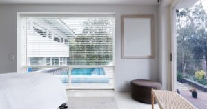 A modern, bright bedroom featuring white walls and a large window with white Venetian blinds that overlooks a swimming pool and lush green trees.