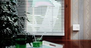 White Venetian blinds covering a window, with a control switch on the wall and a tray of drinks in the foreground.