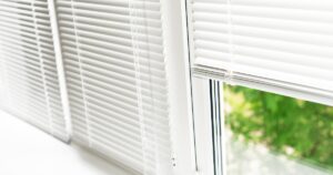 Close-up of white Venetian blinds on a window, partially open to show green foliage outside.