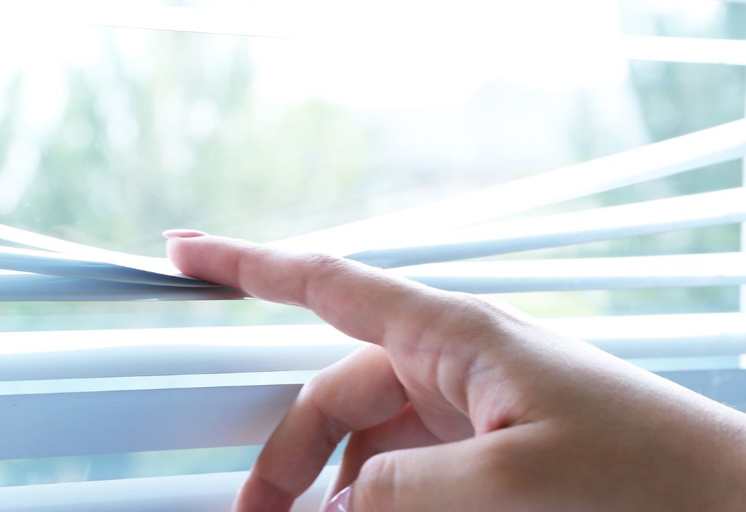 A close-up shot of a person's hand pressing down a slat of white venetian blinds
