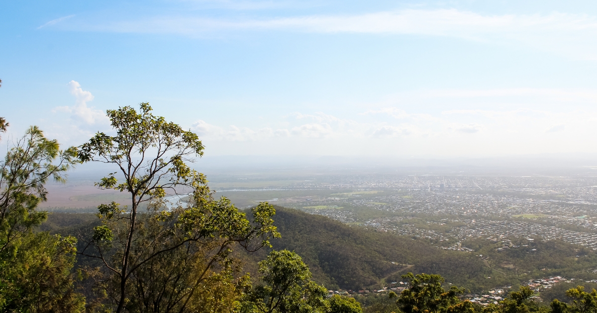 An elevated, wide-angle view of a sprawling coastal city nestled in a valley, framed by lush green trees in the foreground.