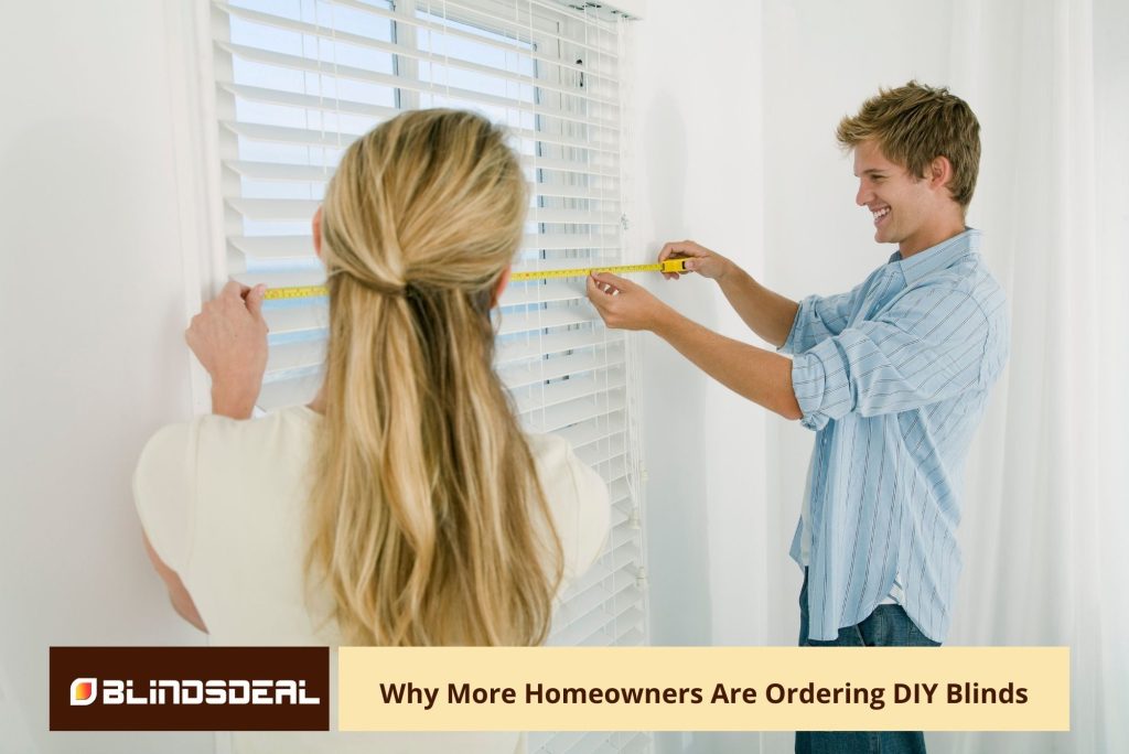 A couple with blonde hair is measuring a window with a yellow tape measure, with their backs to the camera, in front of white horizontal blinds.