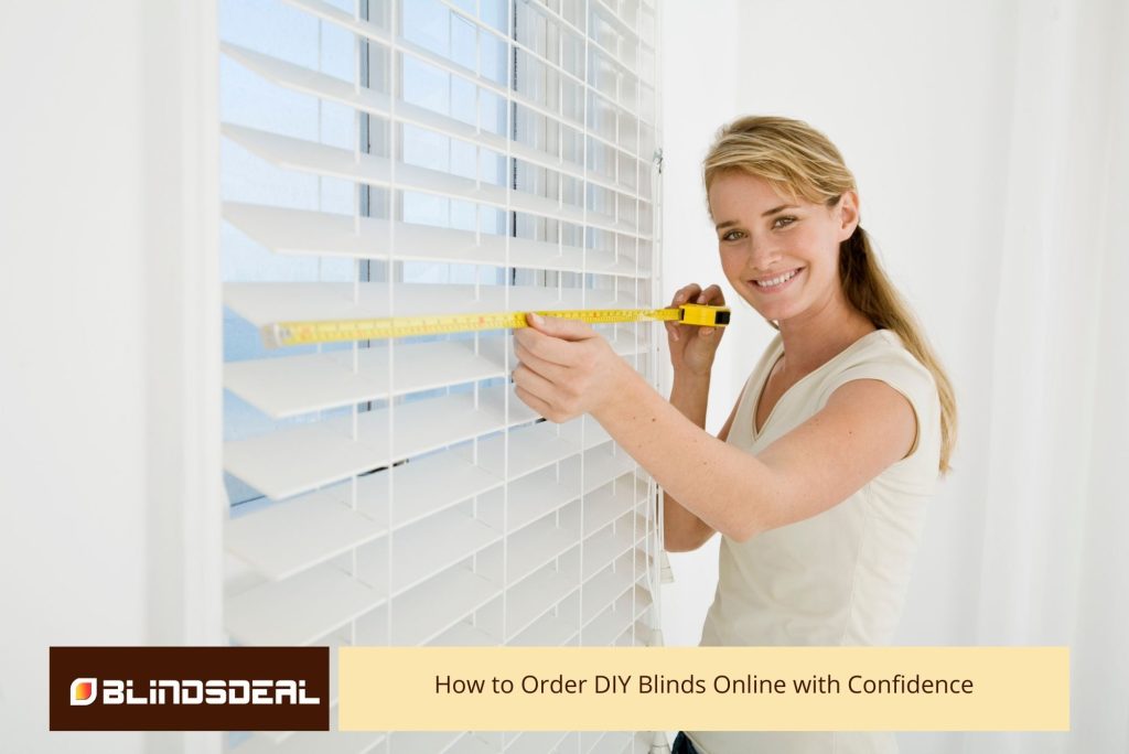 A smiling woman with blonde hair, wearing a white shirt, is measuring the width of a window with a yellow tape measure in front of white horizontal blinds.