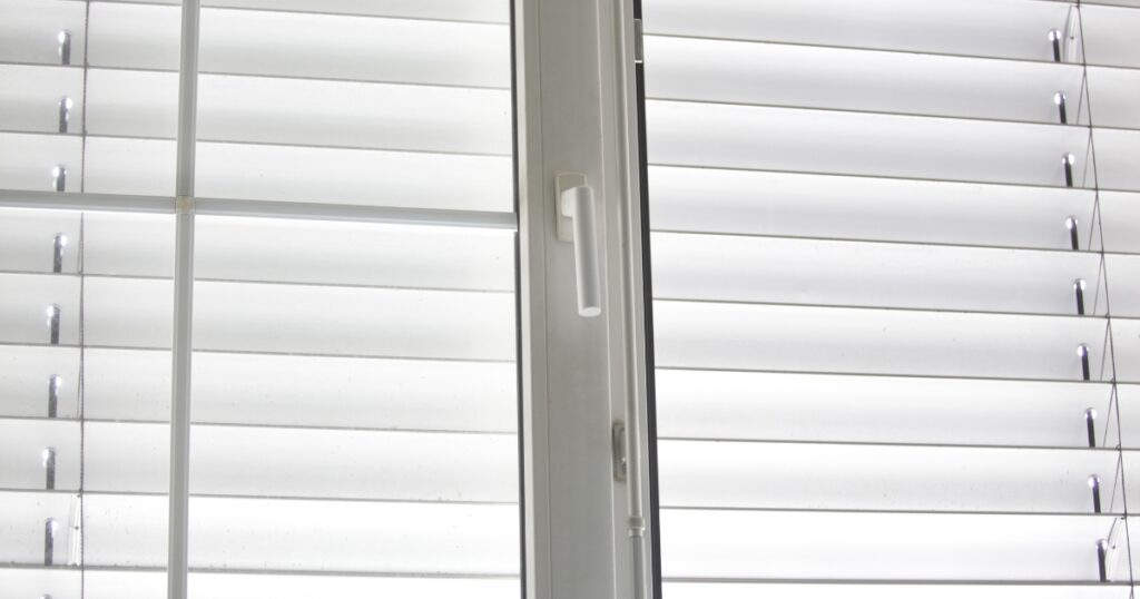 A close-up of a white window frame with a handle, flanked by integrated white horizontal blinds that are partially open