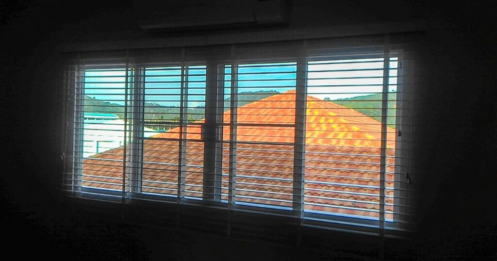 White horizontal blinds partially open to reveal a view of orange tiled roofs