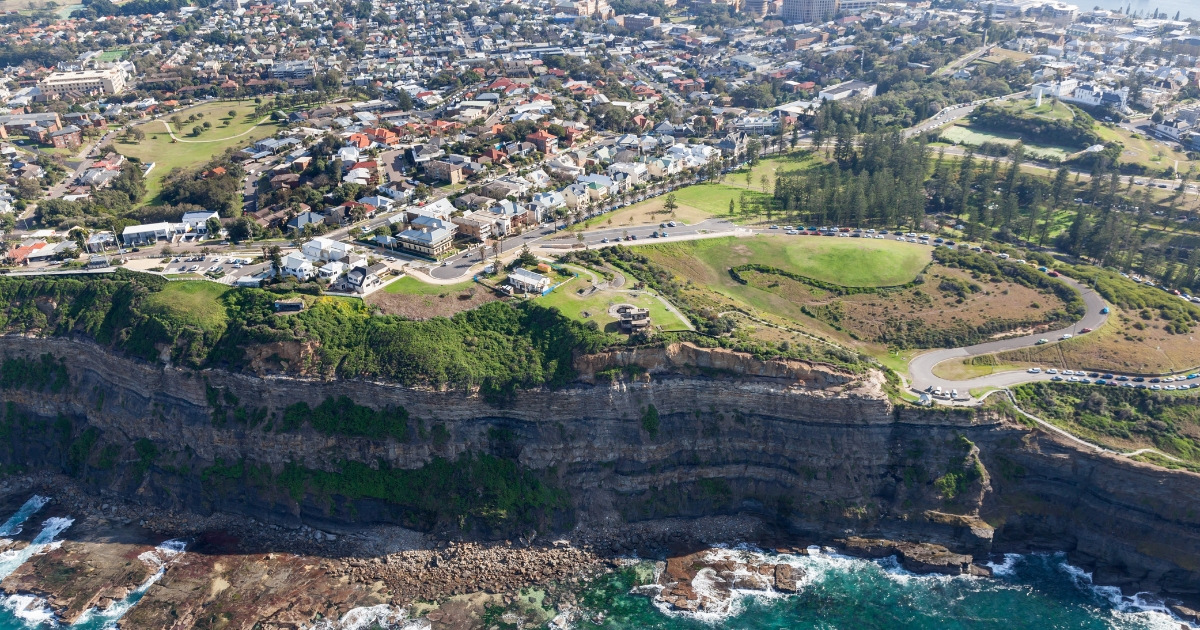 An aerial view of a coastal town featuring steep, rocky cliffs overlooking turquoise ocean waves, with a green park and residential neighborhood stretching inland.