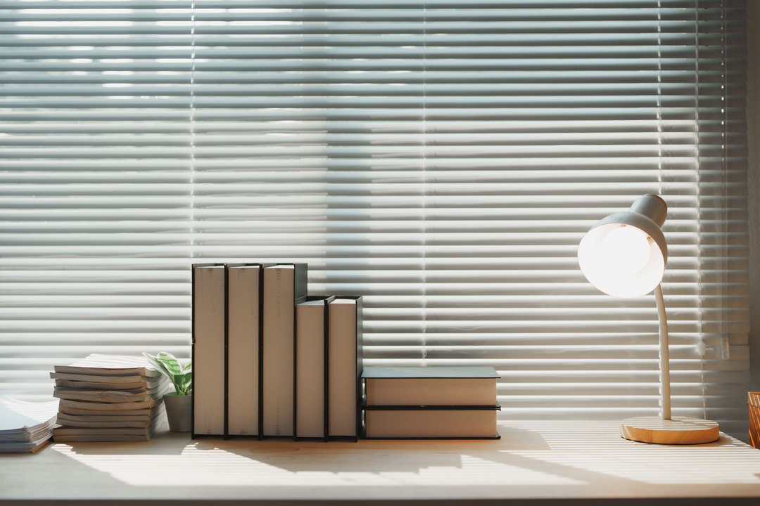 A bright workspace featuring a wooden desk with a white desk lamp and stacks of books against a window with white horizontal blinds.