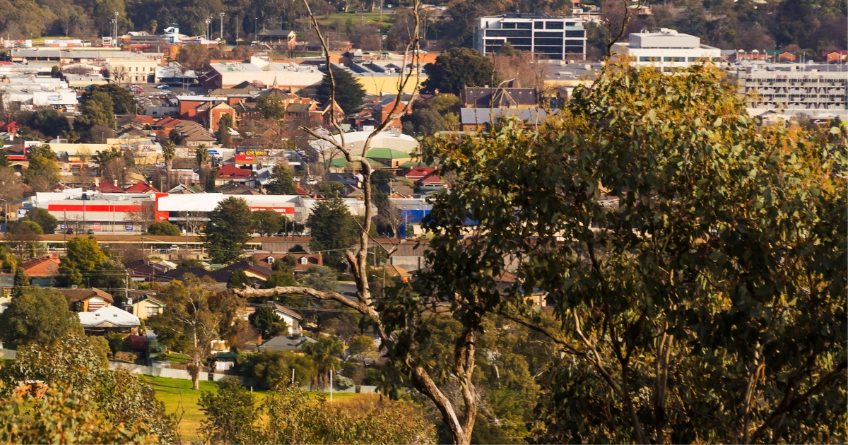 An elevated, wide-angle view of a town nestled among rolling hills, with a mix of residential houses, commercial buildings, and scattered trees