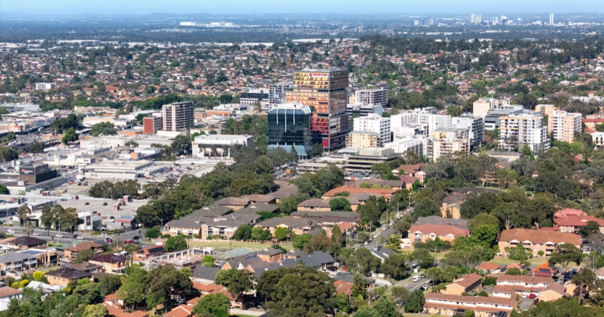 A vast suburban and commercial landscape with a dense layout of houses, green trees, and low-rise apartments surrounding a small cluster of modern office buildings in the centre.
