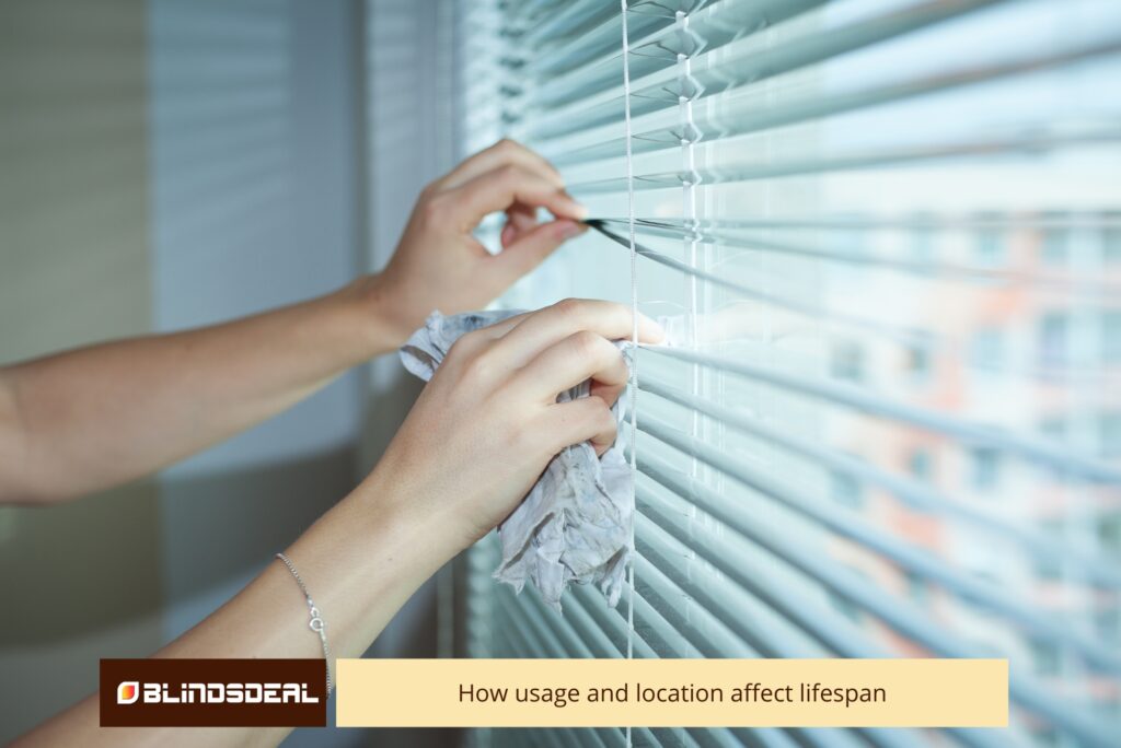 A person's hands wiping down the slats of light-coloured Venetian window blinds with a cloth.