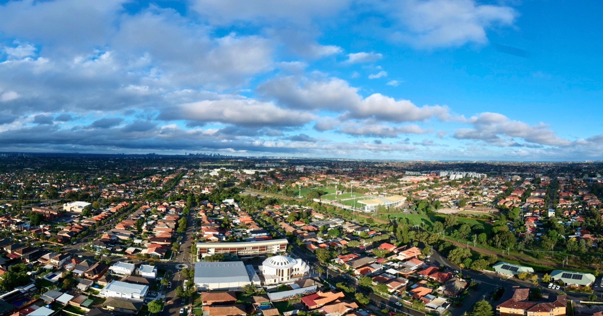 A suburban neighborhood featuring numerous residential houses, trees, and a sports stadium under a blue sky filled with scattered white clouds.