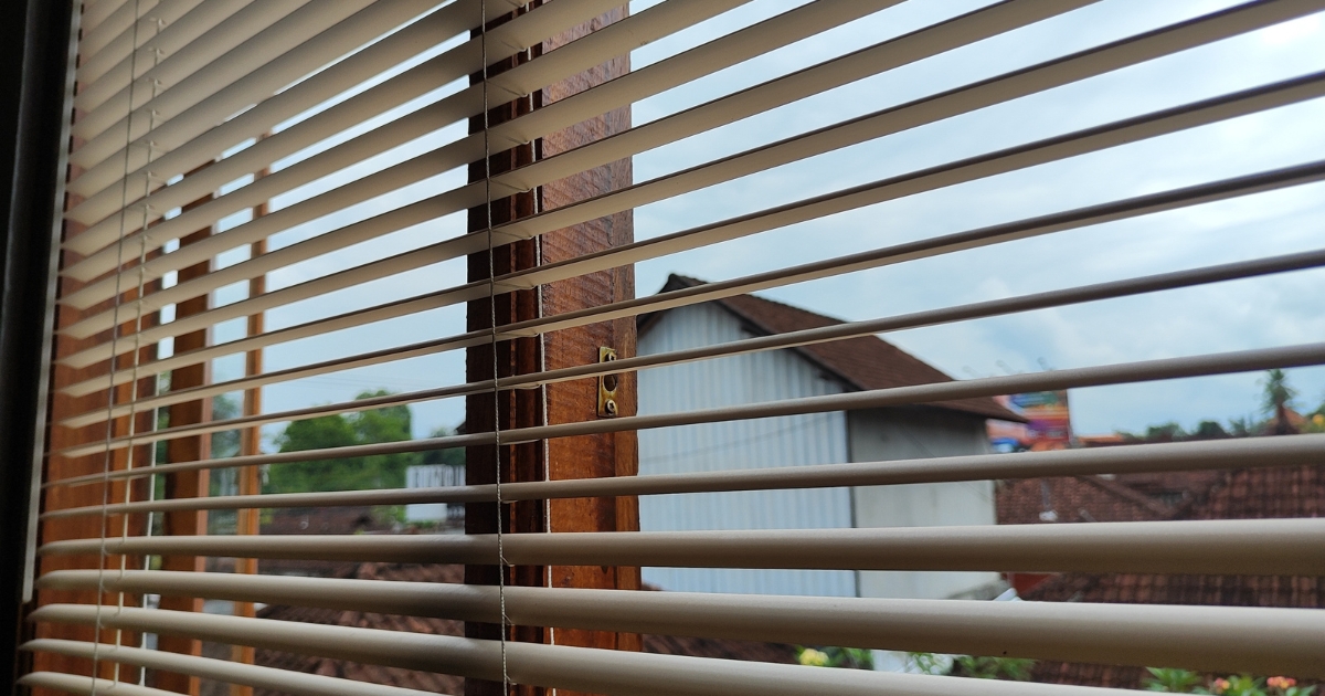 Close-up view of white or light-beige Venetian blinds with slim horizontal slats mounted on a wooden window frame.