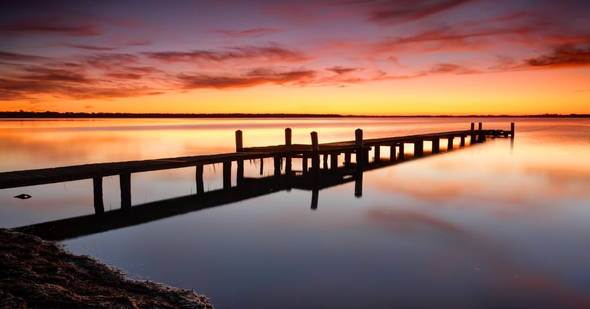 A long wooden jetty extending into calm, reflective water under a dramatic orange and purple sunset sky.