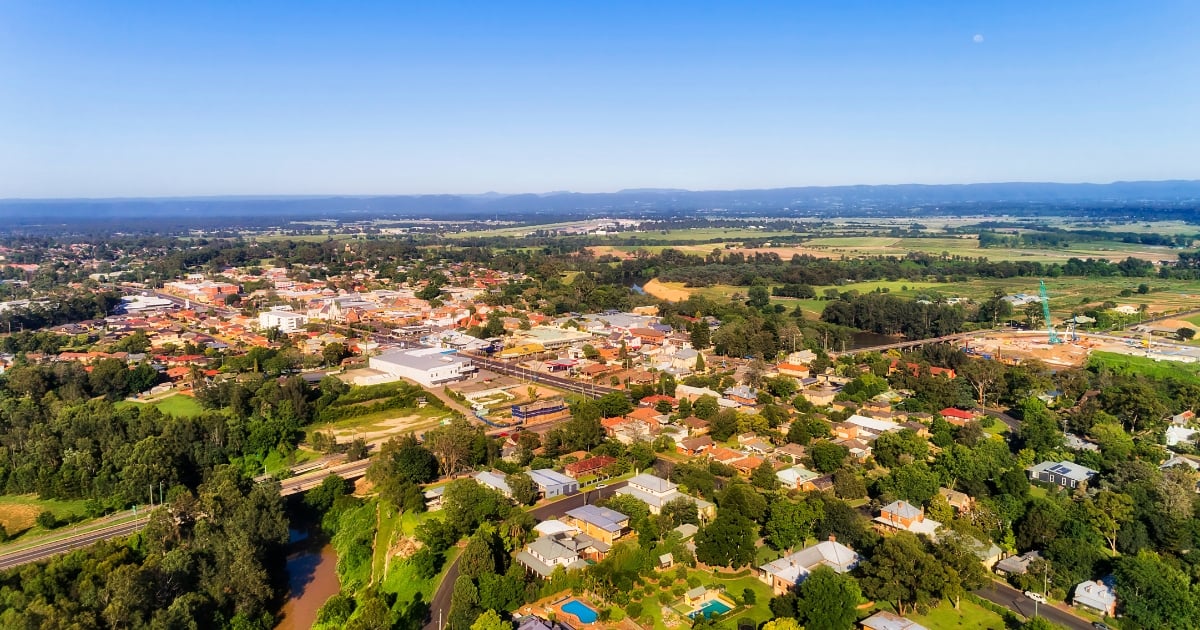 A green suburban town surrounded by fields and forests, with a river winding through the landscape under a clear blue sky.