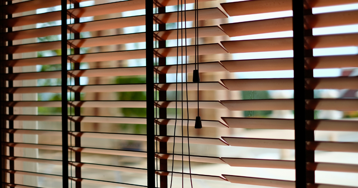 A wooden Venetian blinds with pull cords hanging in the centre, partially open to reveal a softly blurred outdoor background.