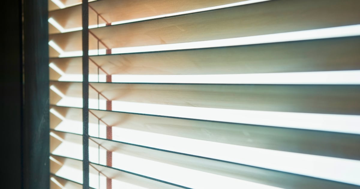 A light-coloured wooden Venetian blinds with sunlight streaming through the open horizontal slats.