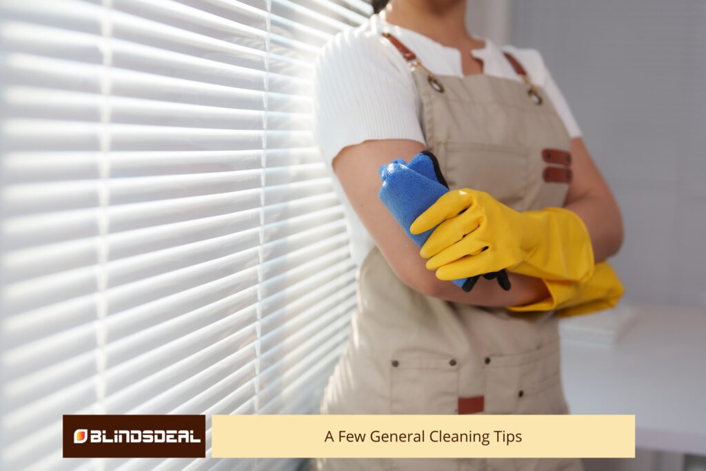 Woman wearing rubber gloves using a feather duster to clean white window blinds.